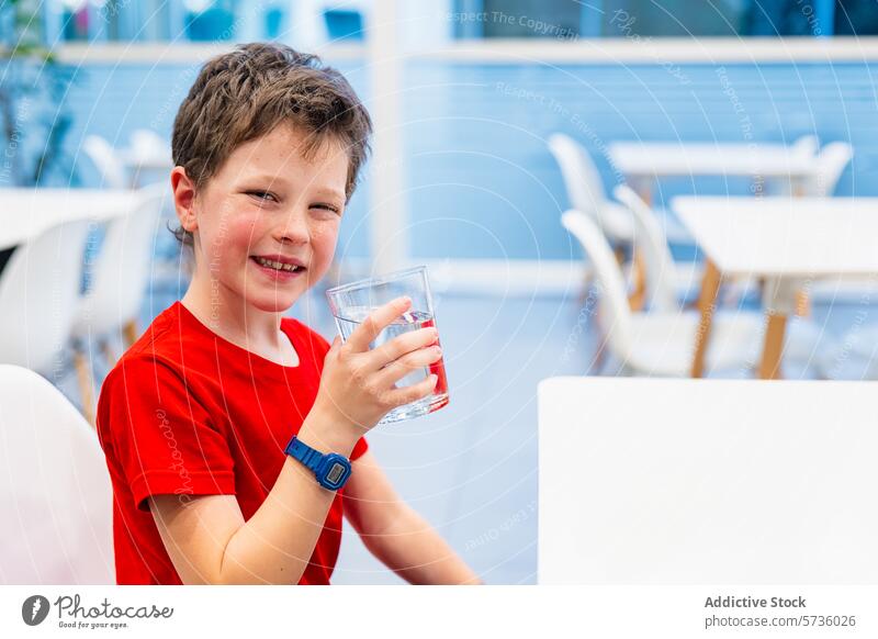 Lächelnder Junge trinkt Wasser in einer hellen Caféumgebung Kind trinken Glas Glück sitzend im Innenbereich rotes Hemd Erfrischung Hydratation Gesundheit