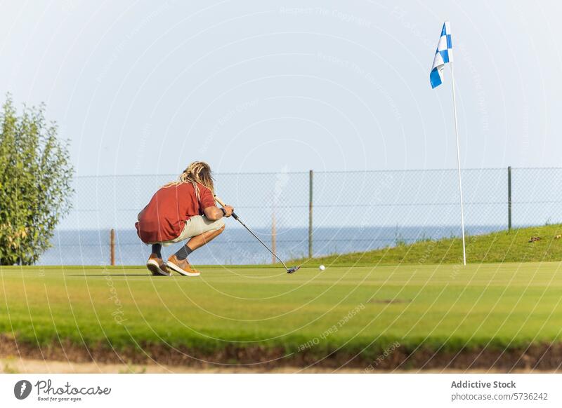 Einzigartiger Golfer mit Dreadlocks bereitet sich auf den Putt vor Rastalocken blond Putten Kurs sonnig hocken Ausrichten Golfschläger Ball grün Fahne Sport