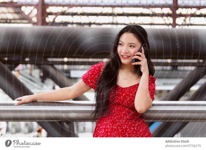 Lächelnde junge Frau, die in einer städtischen Umgebung telefoniert Handy Gespräch rotes Kleid urban Reling Metall verschwommener Hintergrund Großstadt
