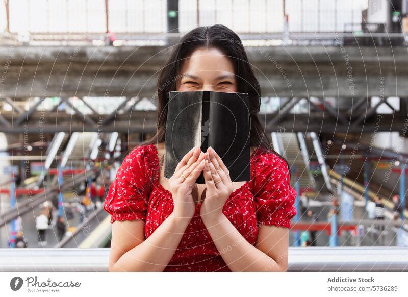 Frau in rotem Kleid verdeckt Gesicht mit Buch am Bahnhof asiatisch deckend spielerisch Tierhaut Lächeln Auge heiter Podest Pendler Porträt beschäftigt reisen
