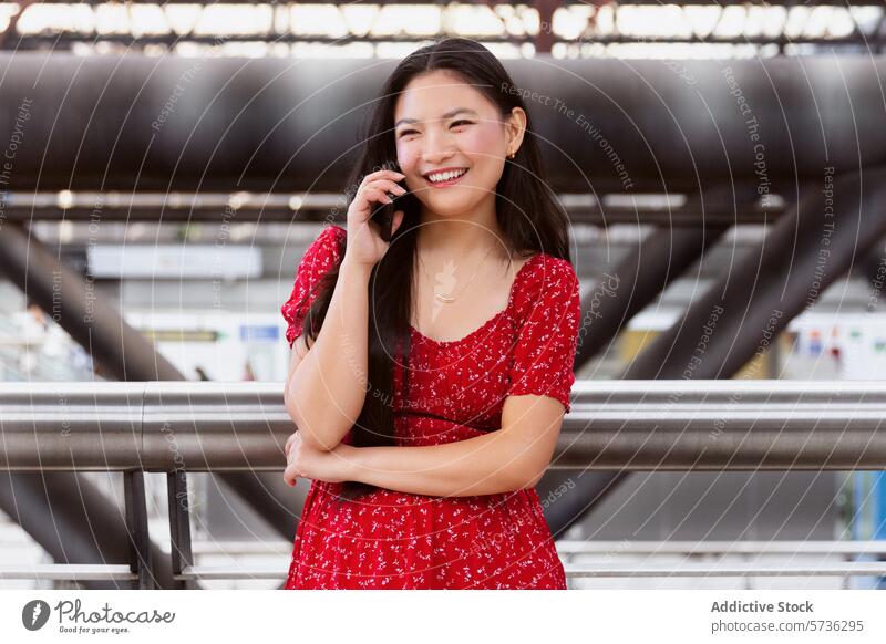 Lächelnde junge Frau, die in einer städtischen Umgebung telefoniert Handy Gespräch rotes Kleid urban Reling Metall verschwommener Hintergrund Großstadt