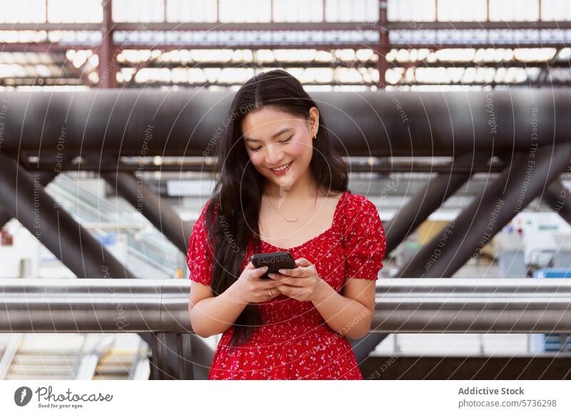 Junge Frau mit Smartphone in einem modernen Bahnhof heiter rotes Kleid jung Architektur Stadtleben Hintergrund Stehen benutzend Telefon Konnektivität Mobile