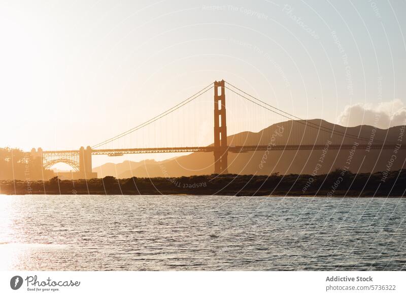 Die untergehende Sonne wirft eine goldene Silhouette der Golden Gate Bridge und schafft eine ruhige Szene über der Bucht mit dunstigem Berghintergrund