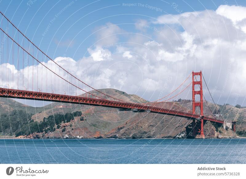 Die Golden Gate Bridge erstreckt sich über blaues Wasser vor einer Kulisse aus Hügeln und flauschigen Wolken und ist das Wahrzeichen von San Francisco. Ikon