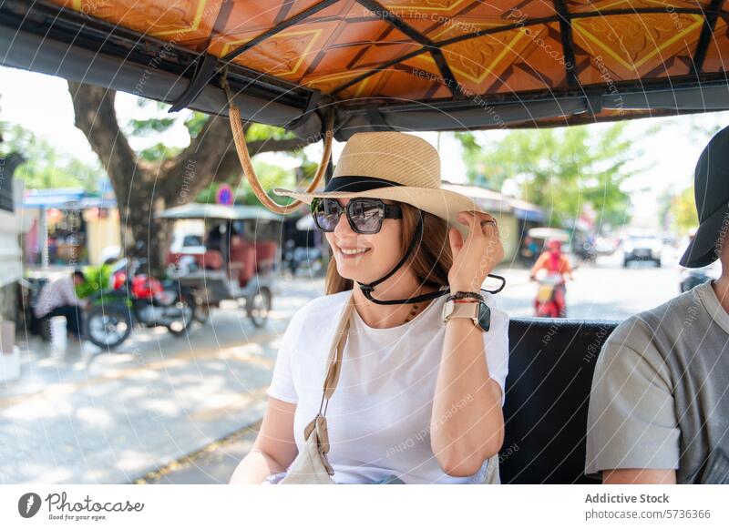 Lächelnde Frau, die eine Tuk-Tuk-Fahrt an einem sonnigen Tag genießt Tuk Tuk Mitfahrgelegenheit heiter Strohhut Sonnenbrille urban reisen Großstadt