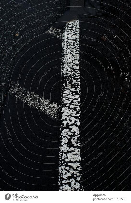 Eine breite weiße Linie auf schwarzem Asphalt. Sie bildet mit weiteren, bereits abgeblassten, schräg verlaufenden Bodenmarkierungen für eine sogenannte Sperrfläche ("Parkverbot") ein runenartiges Zeichen.