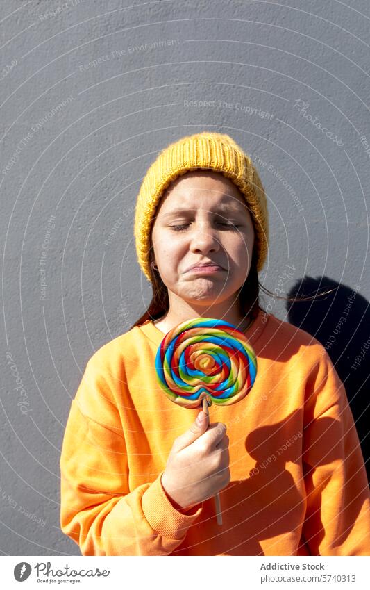 Junge Person mit buntem Lutscher an einem sonnigen Tag jung Lollipop farbenfroh Beanie Sweatshirt orange gelb Stirnrunzeln lässig im Freien unglücklich süß