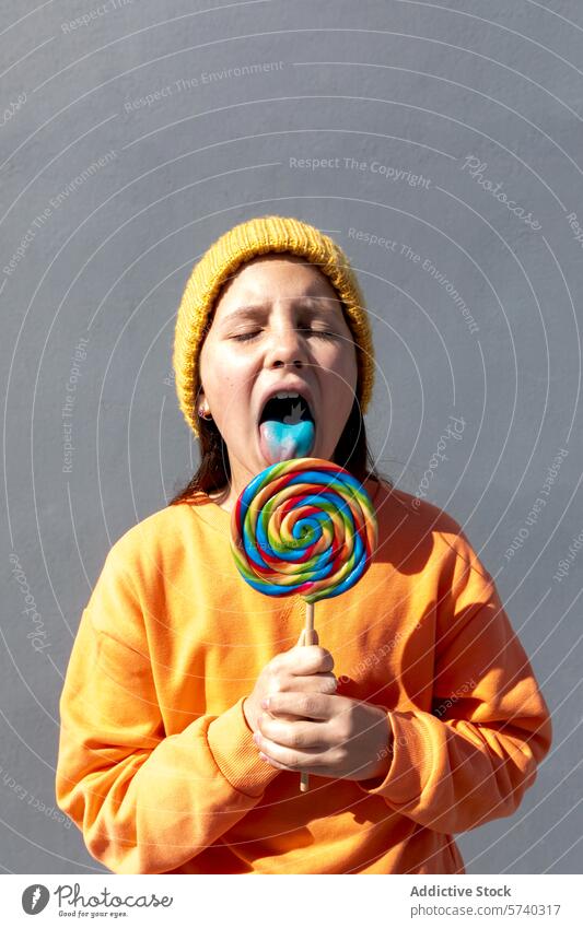 Junge Person genießt einen bunten Lutscher jung einzeln Lollipop farbenfroh orange Sweatshirt gelb Beanie Geschmack groß Verwirbelung Ebene Hintergrund Genuss