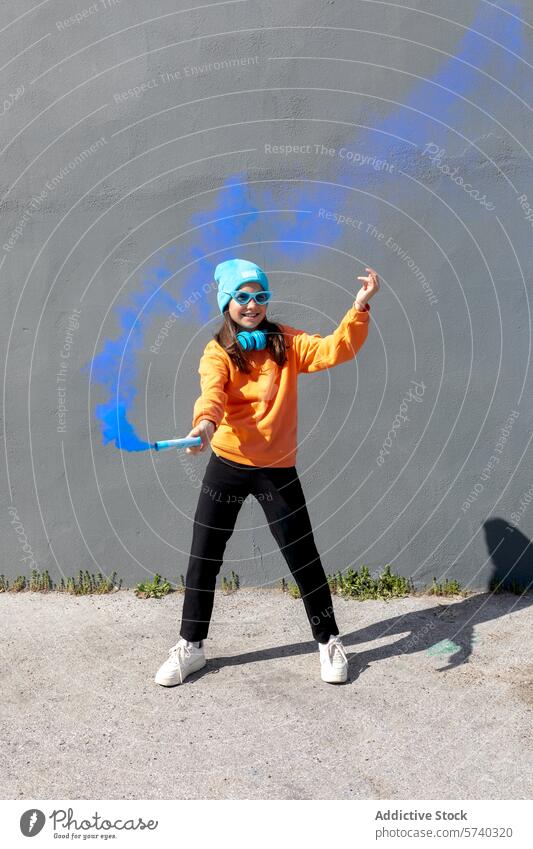 Jugendlicher Geist im lässigen urbanen Stil mit leuchtenden Farben Frau jung heiter Tanzen graue Wand orangefarbenes Sweatshirt blaue Mütze Sonnenbrille