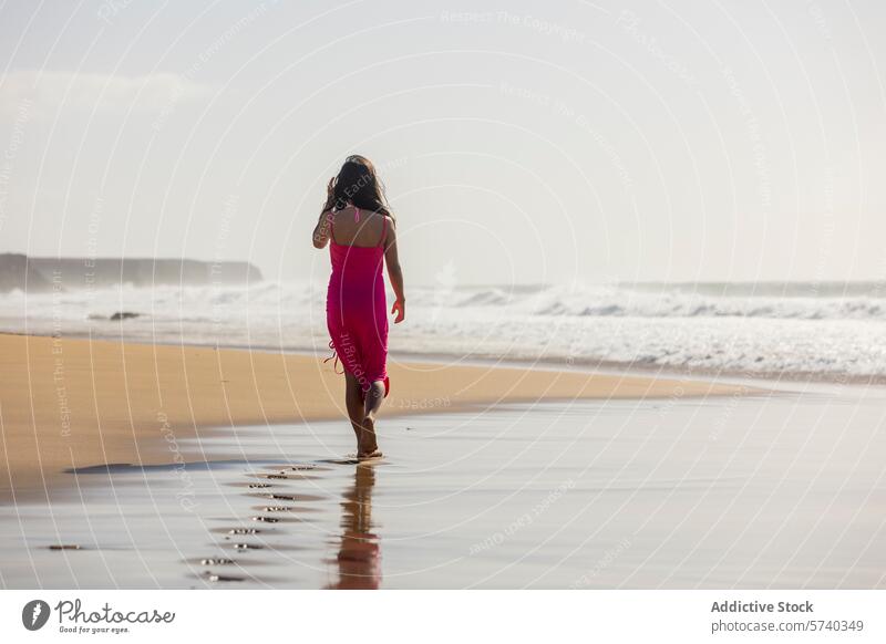 Anonymes junges Mädchen in einem rosa Kleid geht allein am Ufer entlang und hinterlässt Fußspuren im nassen Sand, während sich die Wellen sanft brechen Strand