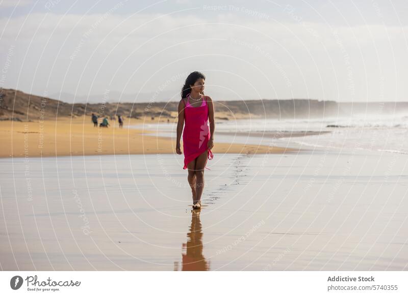 Ein junges Mädchen in einem rosa Kleid geht allein am Ufer entlang und hinterlässt Fußspuren im nassen Sand, während sich die Wellen sanft brechen Strand