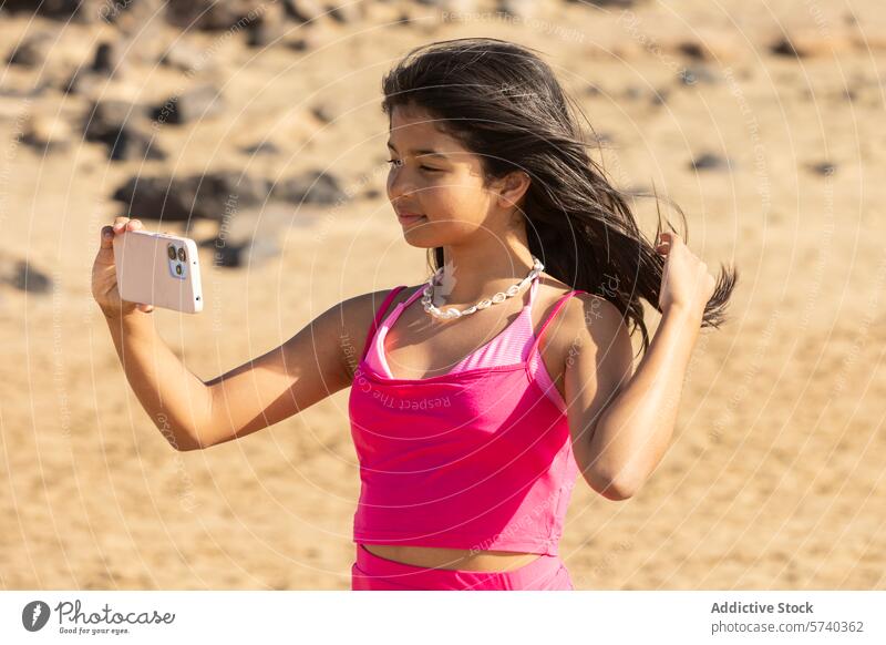 Ein Mädchen in leuchtend rosafarbener Strandkleidung macht selbstbewusst ein Selfie am Sandstrand und fängt so die Essenz eines sonnigen Strandtages ein Telefon