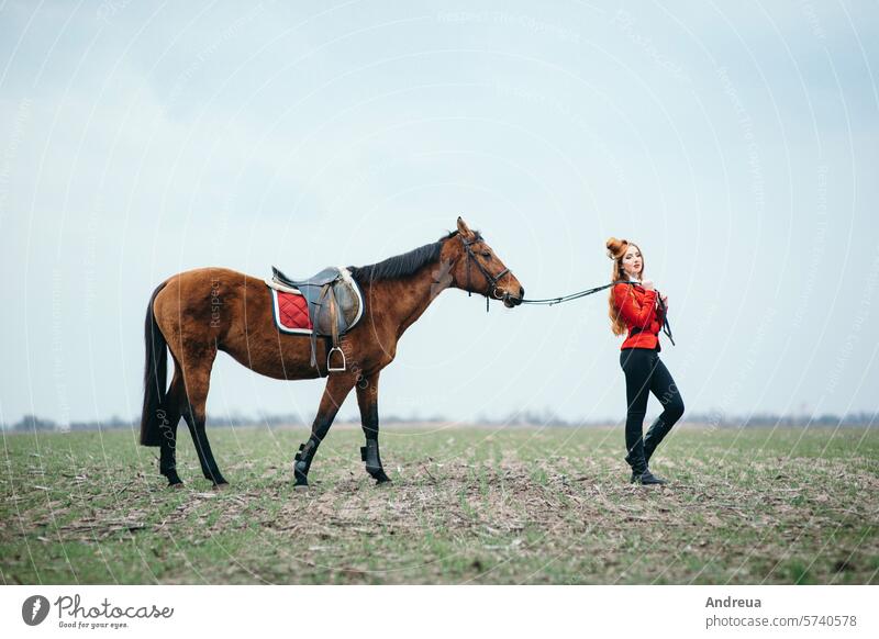 rothaariges Jockey-Mädchen in roter Strickjacke und schwarzen hohen Stiefeln mit einem Pferd Frau Sattel Mitfahrgelegenheit Sommer Natur Reiterin Training