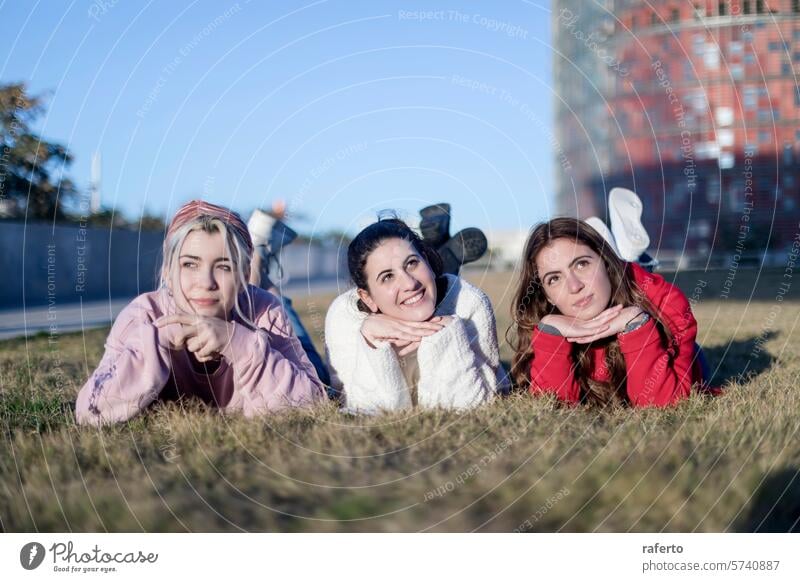 Drei Freunde posieren spielerisch im Gras posierend jung Frauen Klarer Himmel urban Hintergrund liegend Mägen Hände Kinn lässig im Freien Freizeit Tag Glück