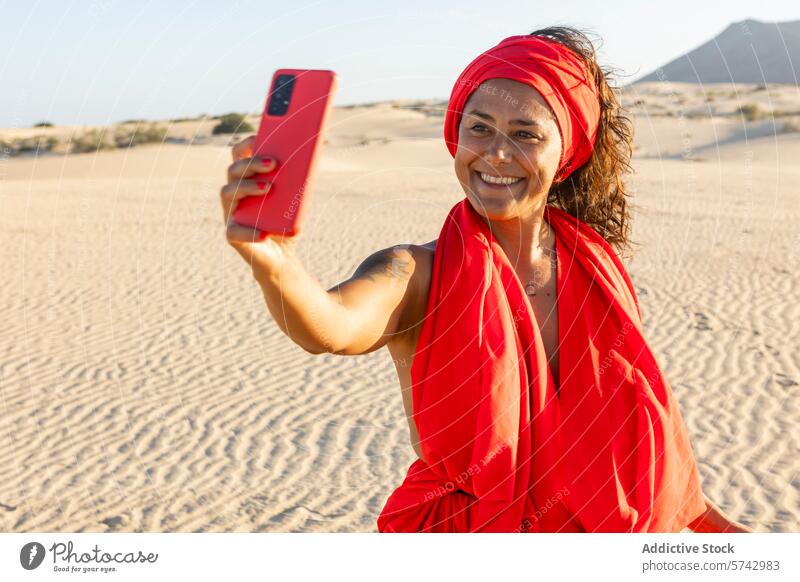 Fröhliche Frau in Rot, die ein Selfie in der Wüste macht rot Kleid wüst Dunes Mobile Telefon Lächeln Fröhlichkeit pulsierend Kopftuch Apparatur Gerät Sand