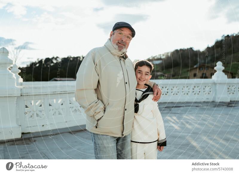 Ein stolzer Großvater mit Mütze umarmt seinen Enkel in Erstkommunionskleidung und genießt den Moment auf einer Terrasse mit Blick auf die Landschaft Umarmen