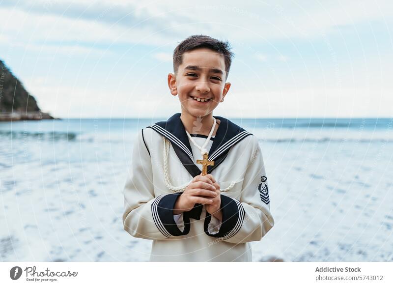 Ein strahlender Junge mit einem Kruzifix in der Hand steht am Meer, gekleidet in seinem Erstkommunions-Matrosenanzug, und verkörpert Freude und Glauben MEER