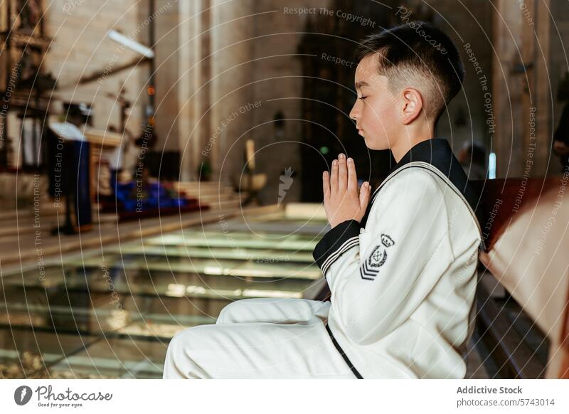 Ein feierlicher Junge sitzt in einer Kirchenbank, die Hände zum Gebet gefaltet, während seiner Erstkommunion, einem heiligen Ritus des Übergangs pew Festakt