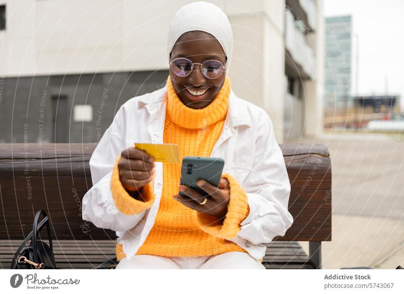 Lächelnde Frau beim Online-Shopping mit Karte und Telefon schwarz Afrikanisch Hijab Brille im Freien Sitzen Bank Kreditkarte Tippen kaufen muslimisch online