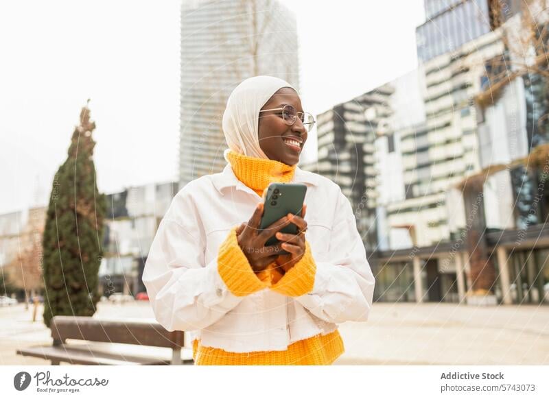 Lächelnde Frau mit Hidschab, die in einer städtischen Umgebung telefoniert Afrikanisch Hijab Smartphone Stadtbild urban heiter Kopftuch Brille modern Gebäude