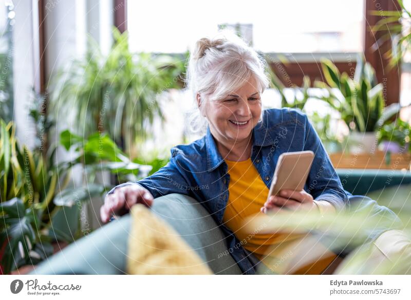 Lächelnde ältere Frau, die ein Smartphone benutzt, während sie zu Hause auf dem Sofa sitzt Menschen lässig Tag Porträt im Innenbereich echte Menschen