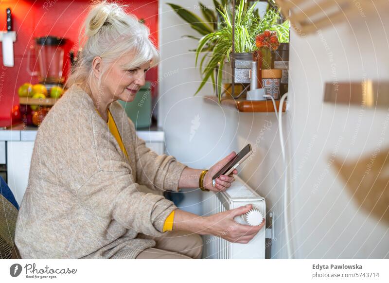 Ältere Frau hält ihr Handy in der Hand, während sie den Thermostat am Heizkörper zu Hause einstellt Heizung Wärme warm Temperatur Komfort Küche Hausheizung
