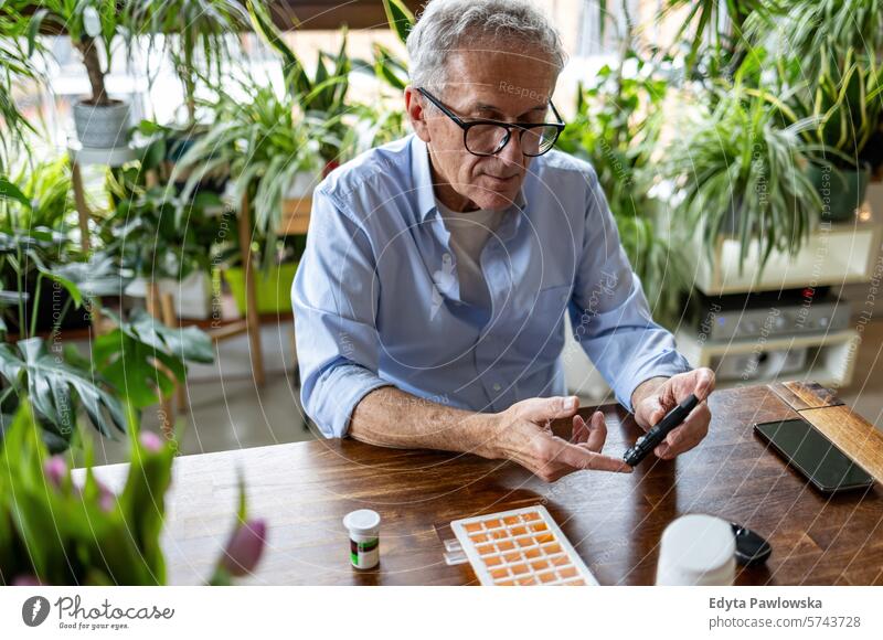 Älterer Mann, der zu Hause sitzend seinen Blutzuckerspiegel überprüft Überprüfung Glaukometer Hände Tisch Gesundheit geduldig Krankheit Sitzen Blutzuckertest