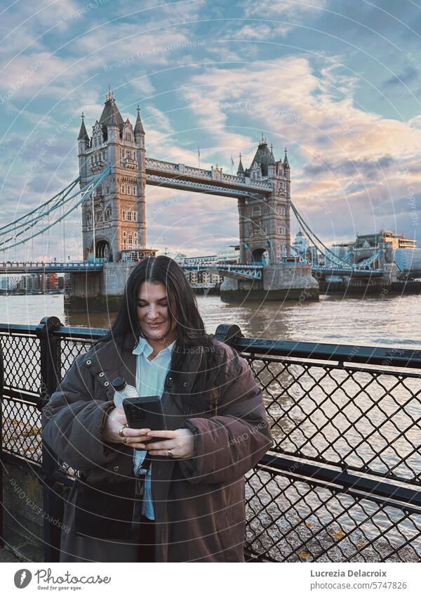 Ein Mädchen steht vor der Tower Bridge in London, schaut auf ihr Handy und lächelt. Lächeln Telefon Tourist reisen Reisen Landschaft Porträt Mode Ausflug Glück