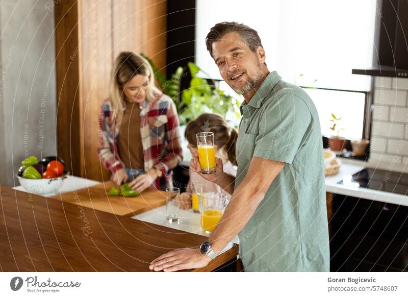 Fröhlicher Mann mit einem Glas Orangensaft, während eine Frau und ein Kind in einer gemütlichen Küche Essen zubereiten Familie Lächeln Essenszubereitung