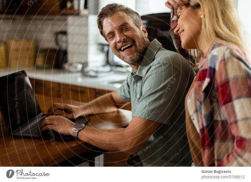 Ein fröhliches Paar genießt einen unbeschwerten Moment in seiner sonnigen Küche, während es am Laptop arbeitet und ein gesundes Frühstück zu sich nimmt Lachen