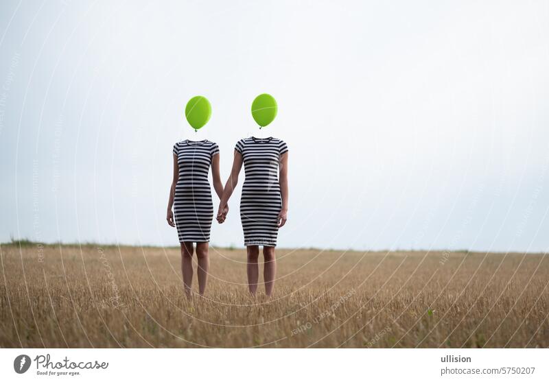 zwei glückliche, sexy junge Frauen in schwarz-weiß gestreiften Kleidern, Freundinnen, halten sich an den Händen, die Köpfe durch grüne Luftballons ersetzt, stehen Hand in Hand auf einem abgeernteten Maisfeld