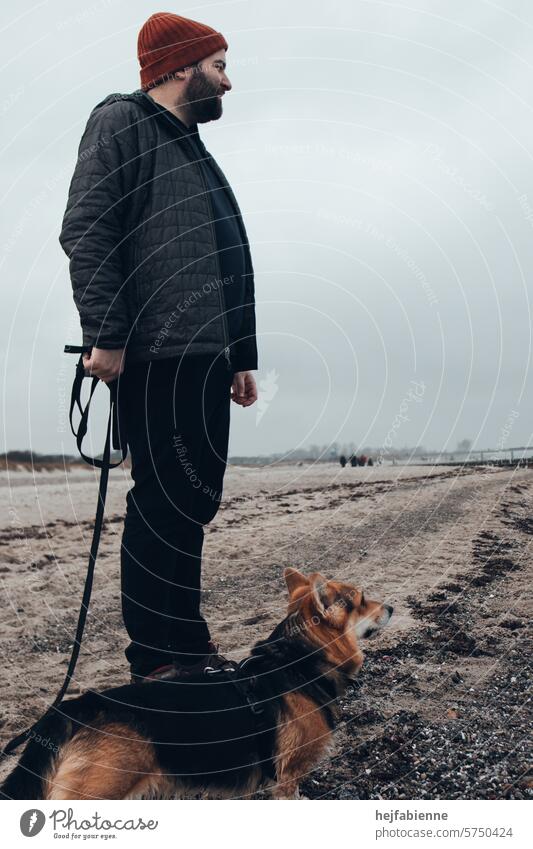 Neblige Gassirunde am Ostsee Hundestrand | Herrchen und Corgi blicken aufs Meer | Männlicher Hundehalter mit Vollbart und roter Mütze Haustier Gassi gehen