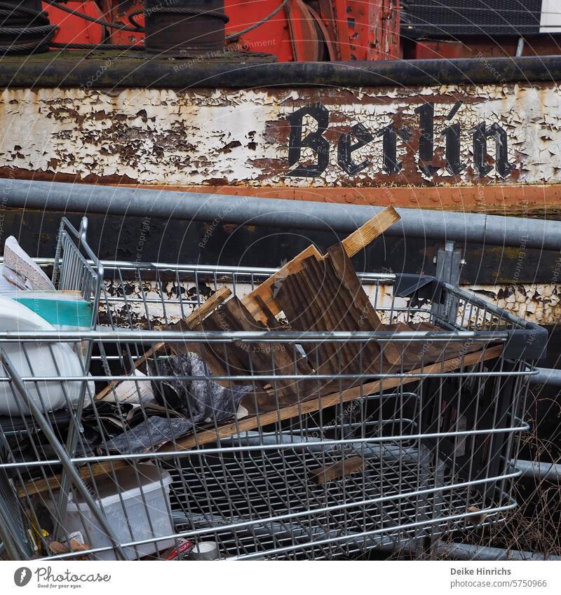 Einkaufswagen mit Müll gestrandet vor altem Kahn mit Aufschrift Berlin berlinerwasser Großstadt urban Schiff rostig mobilität Stiefel einkaufen abgestellt