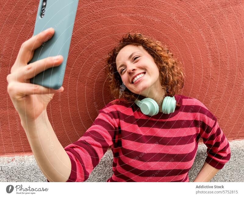 Fröhliche Rothaarige machen ein Selfie mit Smartphone Frau Rotschopf Kopfhörer Glück heiter Technik & Technologie soziale Netzwerke rote Wand Jugend Lifestyle