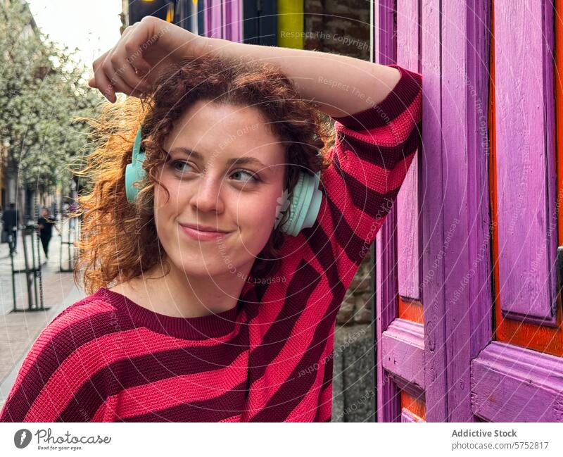 Lächelnde junge rothaarige Frau mit Kopfhörern, die an einer lila Tür lehnt Musik krause Haare gestreift Hemd purpur pulsierend Großstadt Straße Freizeit lässig