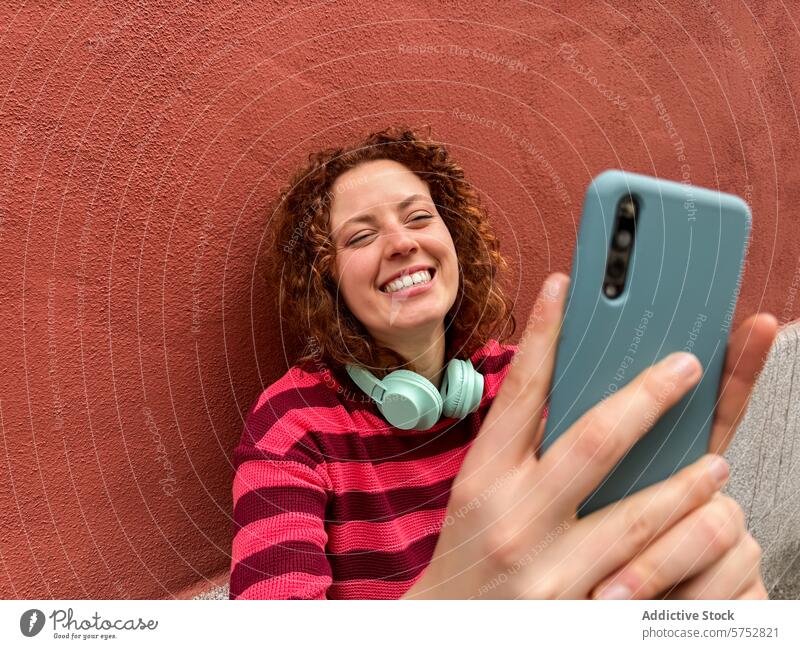 Junge rothaarige Frau genießt einen Videoanruf auf dem Smartphone Video-Chat Lächeln roter Hintergrund jung Erwachsener Spaß Streifenpullover
