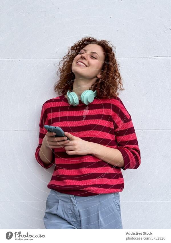 Lächelnde junge rothaarige Frau, die mit Kopfhörern Musik genießt krause Haare Smartphone weißer Hintergrund Genuss Freizeit Fröhlichkeit Streifenpullover Mode