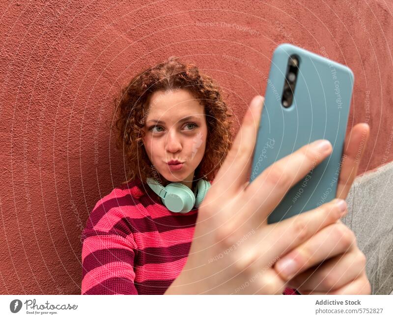 Junge rothaarige Frau, die ein Selfie mit einem verspielten Ausdruck macht Smartphone spielerisch krause Haare rote Wand Falten Lippen Beteiligung lässig Jugend