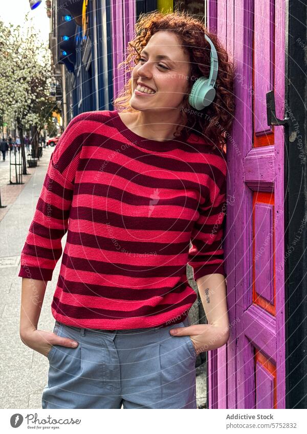 Lächelnde junge rothaarige Frau genießt Musik in städtischer Umgebung Kopfhörer urban Tür farbenfroh heiter krause Haare Streifenpullover Lehnen Genuss
