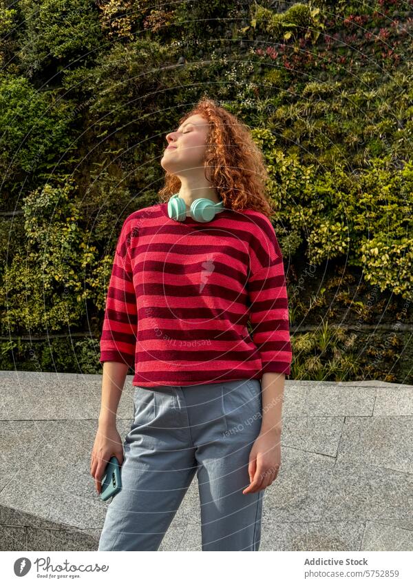 Junge rothaarige Frau genießt den Sonnenschein in Freizeitkleidung jung krause Haare Lächeln Sonnenlicht Kopfhörer Smartphone lässig Erholung friedlich Genuss