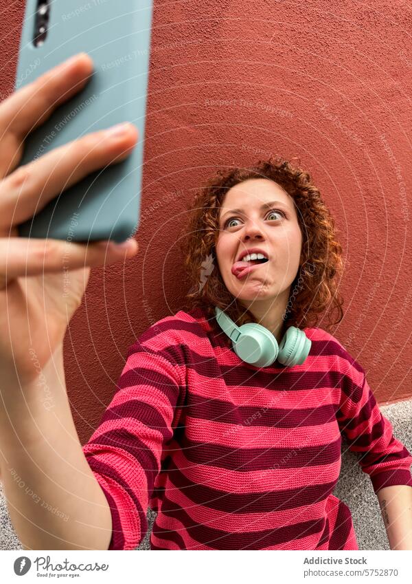 Rothaarige Frau macht ein lustiges Gesicht, während sie ein Selfie macht spielerisch Grimasse Smartphone rote Wand krause Haare Kopfhörer Ausdruck Humor lässig