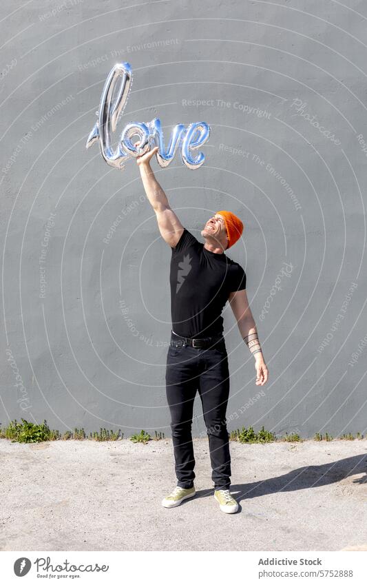 Mann hält einen Liebesballon mit freudigem Ausdruck Luftballon Glück glänzend Halt schwarzes Outfit orangefarbene Mütze Wand Feier romantisch Konzept Zuneigung