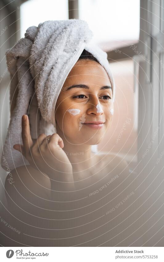 Frau trägt Gesichtscreme für die Hautpflege auf Feuchtigkeitscreme Schönheit Routine Handtuch Kopf anwendend Gelassenheit Fenster Morgen Wellness Pflege