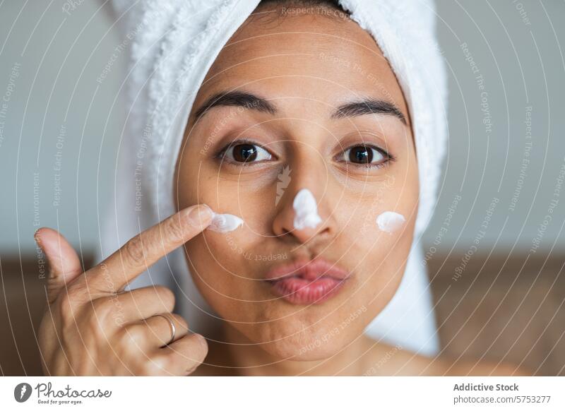 Verspielte Frau trägt Feuchtigkeitscreme auf ihr Gesicht auf Handtuch Headwrap Hautpflege Gesichtscreme spielerisch Ausdruck Schönheit Routine Pflege Gesundheit
