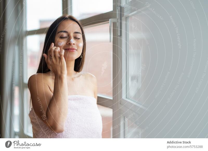 Frau trägt am Fenster Gesichtscreme auf Hautpflege Gesichtsbehandlung Sahne Feuchtigkeitscreme Schönheit Routine anwendend Pflege Handtuch Wellness hell Hygiene