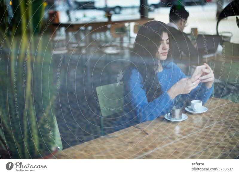 Frau schreibt in einem Café eine SMS Texten Smartphone blau Pullover Kaffee Tasse Untertasse Sitzen Fenster Reflexion & Spiegelung entspannt allein