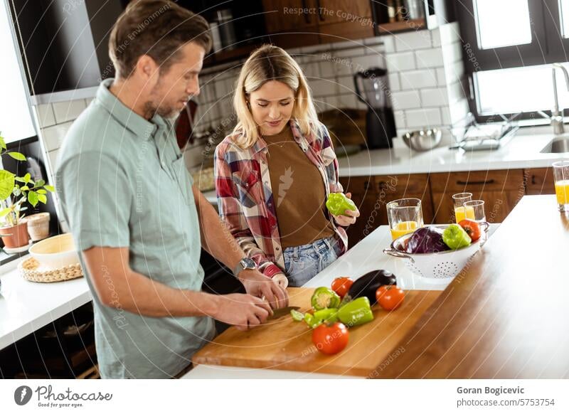Ein lächelnder Mann und eine lächelnde Frau, die auf einer Kücheninsel frisches Gemüse hacken und gemeinsam eine gesunde Kochaktivität genießen Paprikaschoten