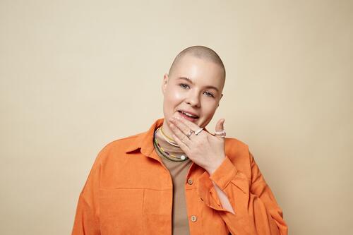 Porträt einer selbstbewussten jungen Frau mit Glatze und leuchtend orangefarbener Jacke im Studio kahl Mädchen gen z Haarausfall Alopezie Mund Lippen lutschen