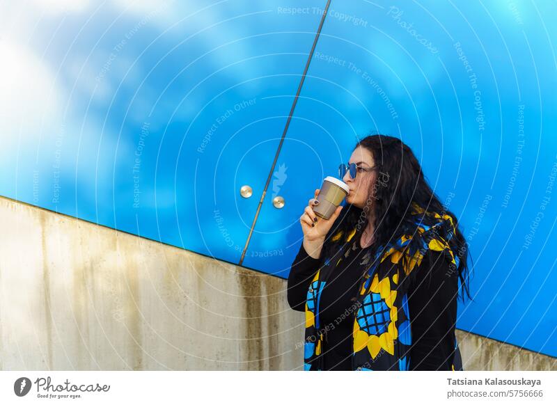 Schöne junge brünette Frau mit langen lockigen Haaren, gekleidet in einer schwarz-blau-gelben Weste, trinkt Kaffee aus einem Pappbecher vor einem blauen Wandhintergrund