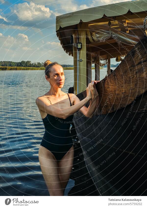 Eine junge, schlanke, attraktive Frau in einem einteiligen Badeanzug entspannt sich am See und lehnt an einem hölzernen Passagierschiff Stehen Wasser alt